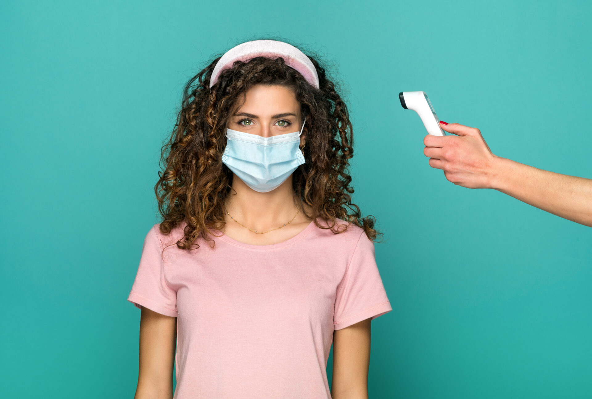 Young woman in protective face mask during the Covid-19 or coronavirus pandemic having her temperature monitored on a digital thermometer over a blue studio background