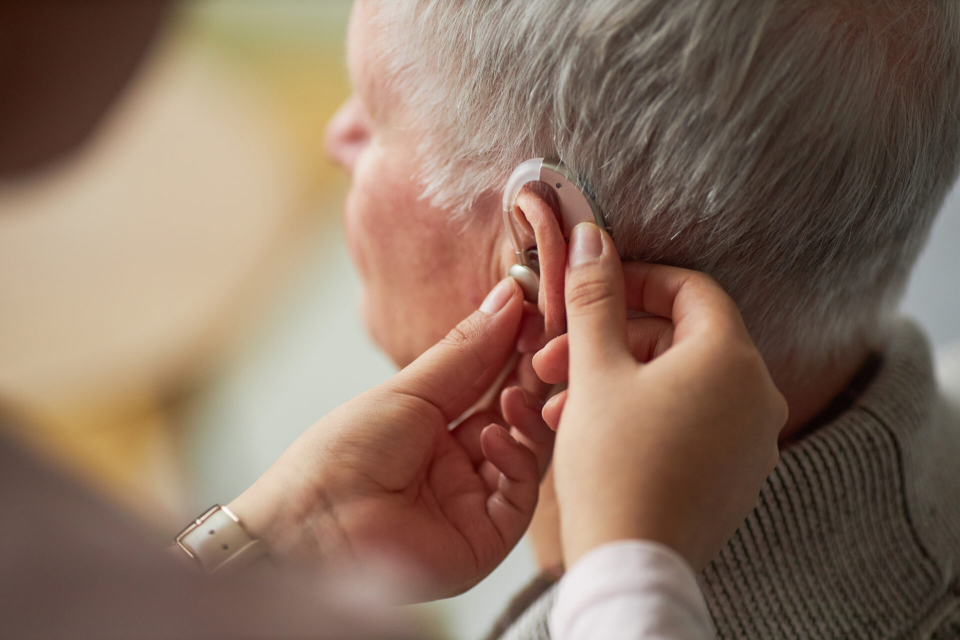 Close-up of care worker helping a senior man with deafness to wear a hearing device on his ear.